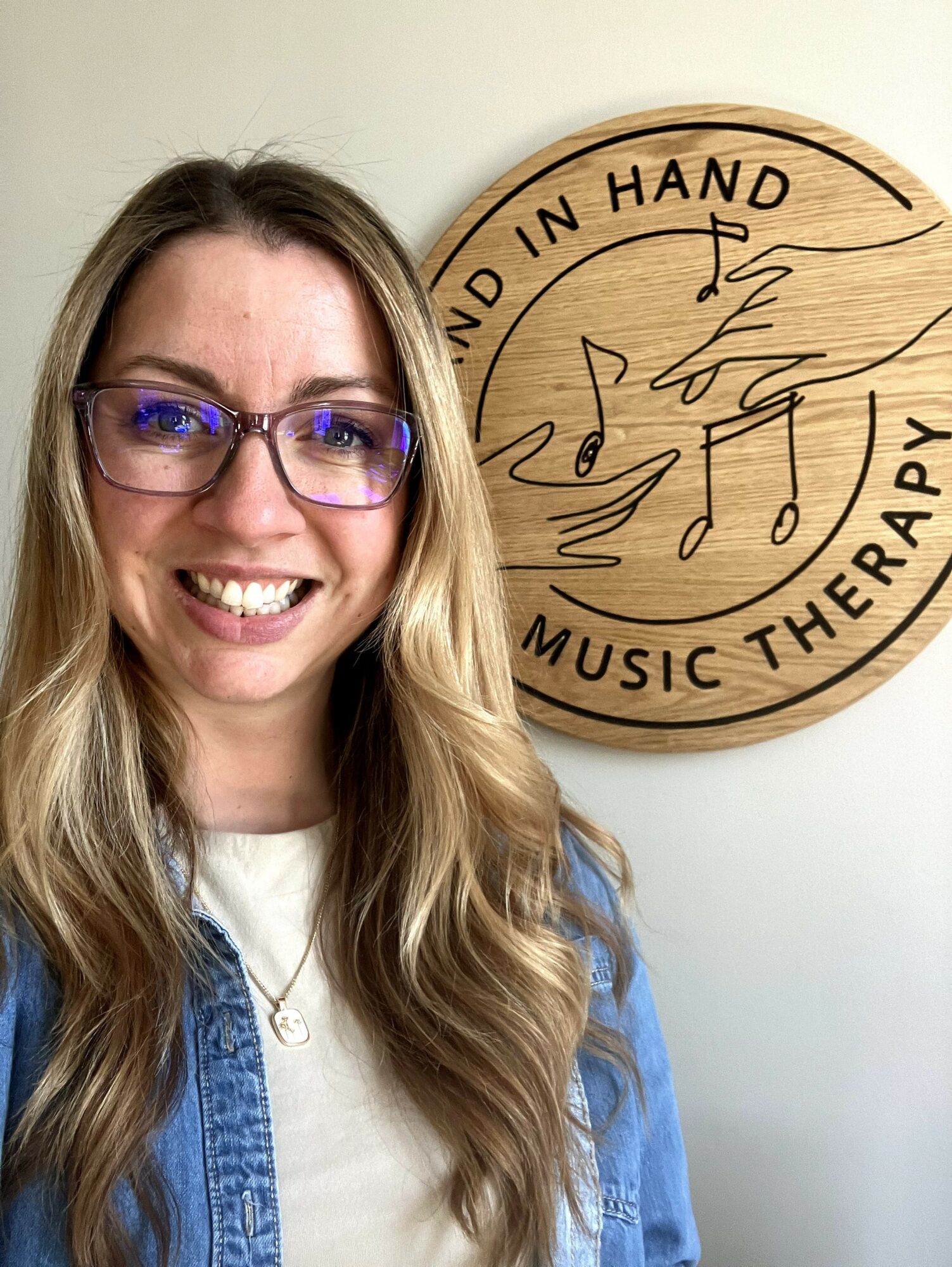 Woman with glasses smiling, long wavy hair, standing next to a round wooden sign with musical notes and text.