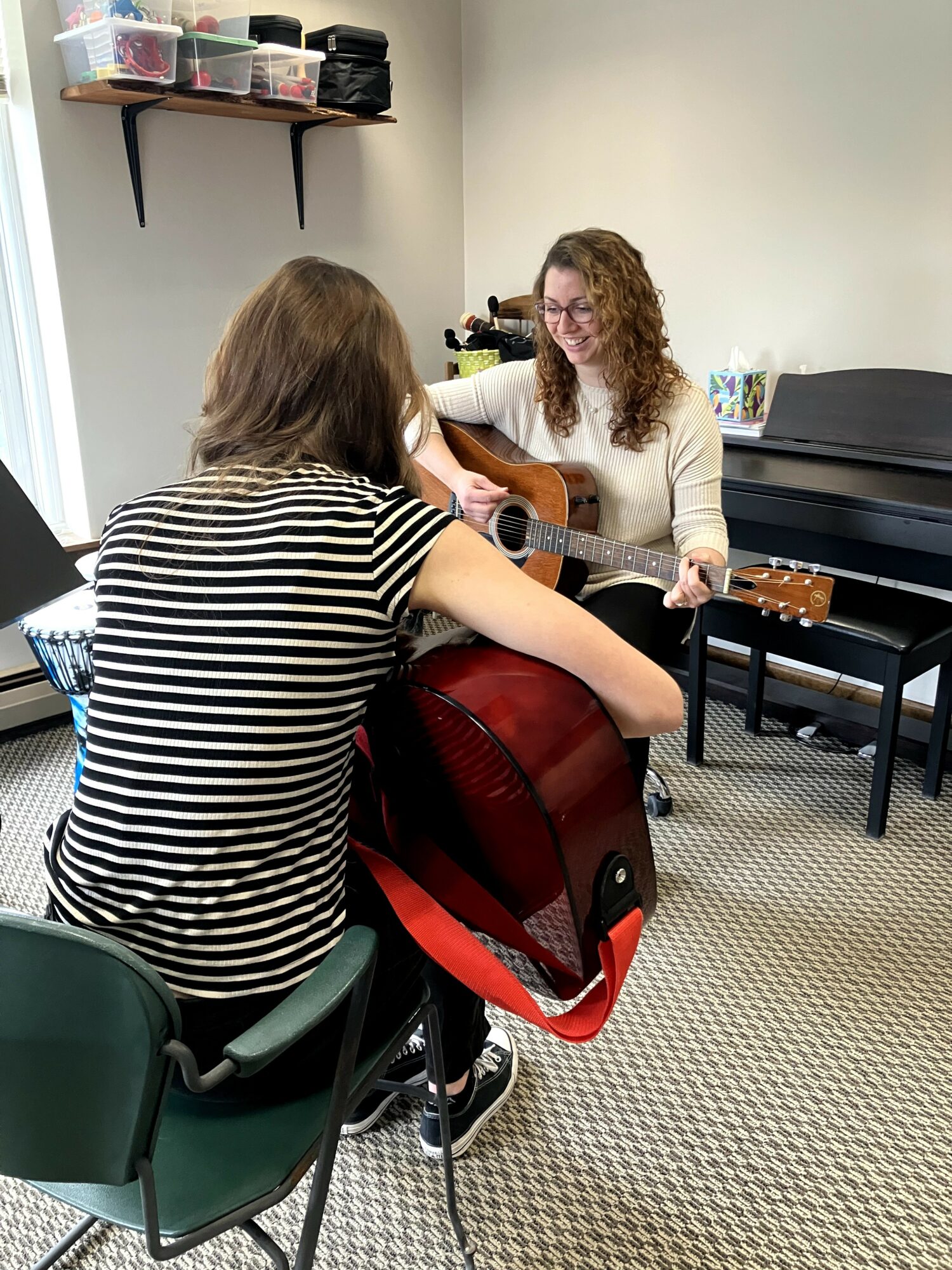 Two women sitting in a music room, one playing guitar, the other watching, with a piano and shelves in the background.