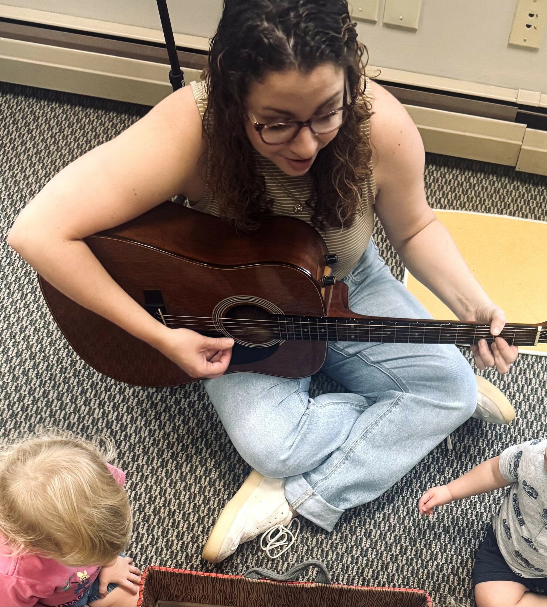 Woman sitting on carpeted floor playing guitar with two children nearby, one child in pink and one in striped shirt.