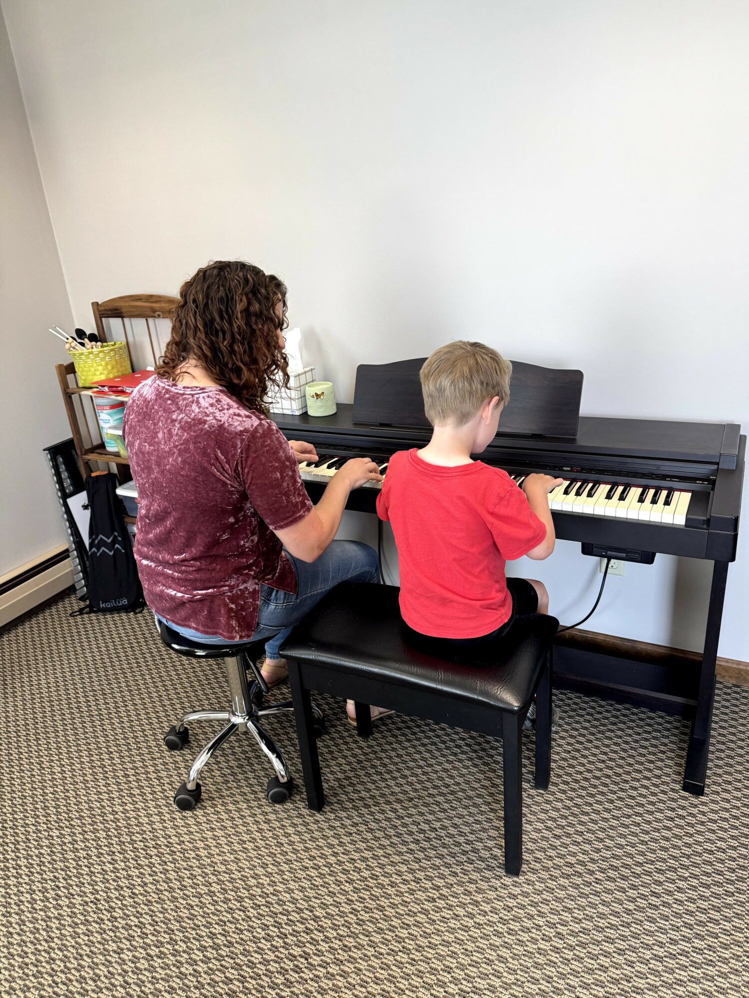 Woman and child sitting at a black digital piano, facing away, in a room with white walls and beige carpet.