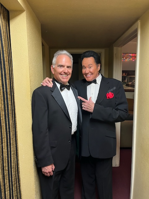 Two men in tuxedos smiling and posing together in a hallway, one with hand on the other's shoulder, both wearing bow ties.