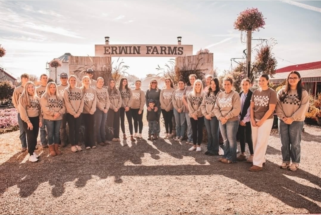 Group of people standing outdoors in front of a sign that reads 'ERWIN FARMS', with trees and a building in the background.