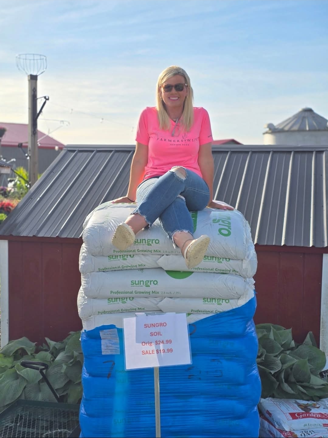 Woman sitting on stacked bags outdoors, smiling, wearing sunglasses and a pink shirt, with a building and sky in background.