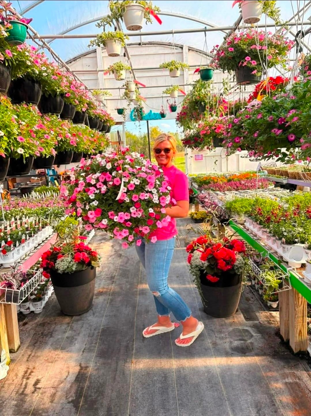 Person in pink shirt and jeans holding a large pink and white flower arrangement in a greenhouse filled with hanging and potted flowers.