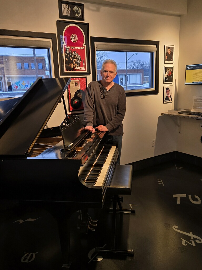 Older man with white hair standing next to a grand piano in a room with framed pictures and windows.