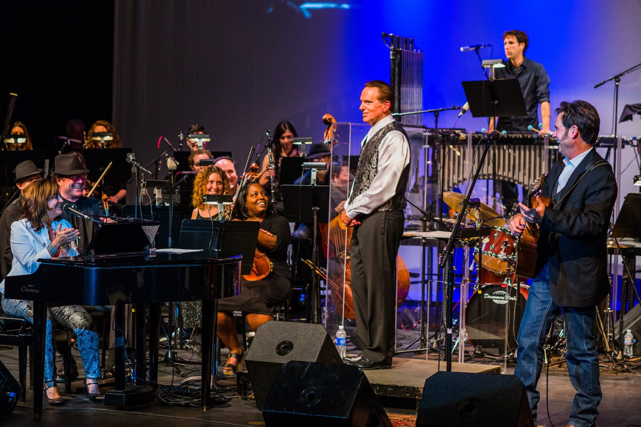 Orchestra performing on stage with musicians playing various instruments, conductor, and a percussionist in the background, stage lighting, and music stands.