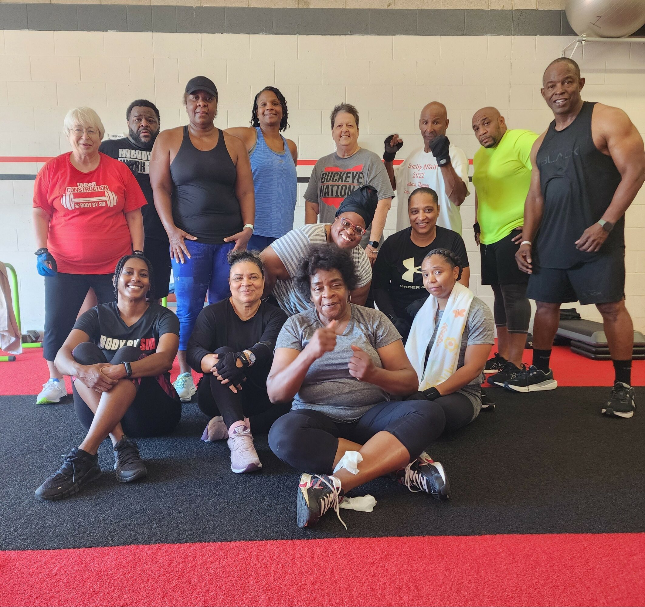 Group of fifteen people in workout attire posing indoors on a gym floor, some smiling and making gestures.