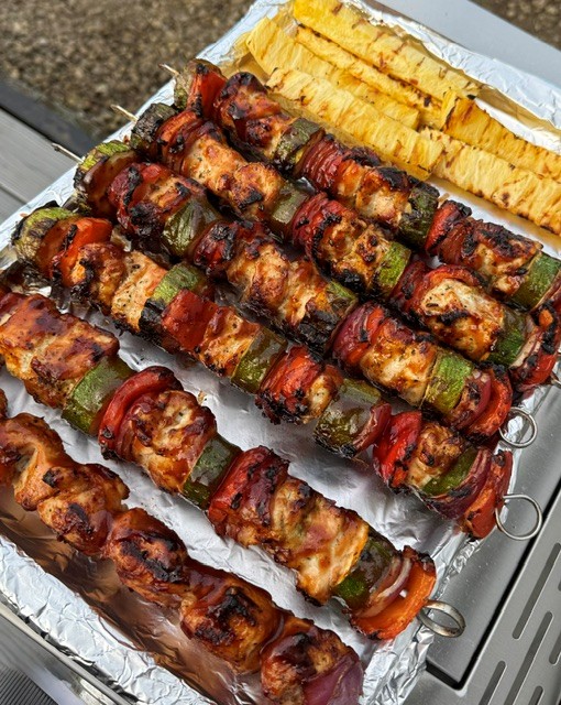 Barbecue chicken and vegetable skewers with grilled pineapple slices on foil-lined tray.