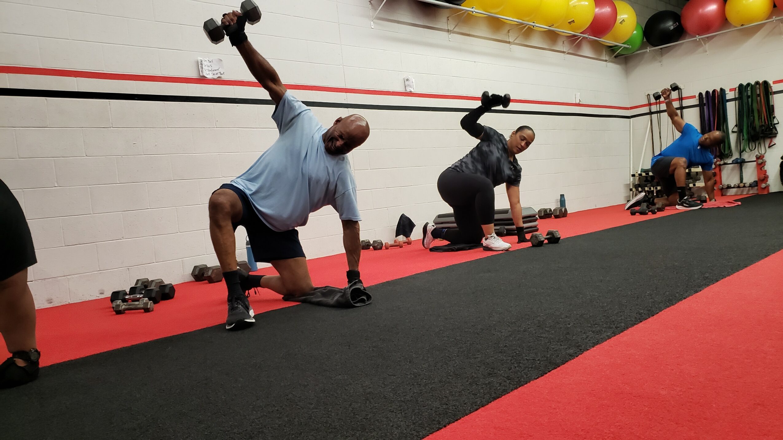 Three people exercising with dumbbells in a gym, one person lifting weights overhead, others on the floor, red and black flooring.