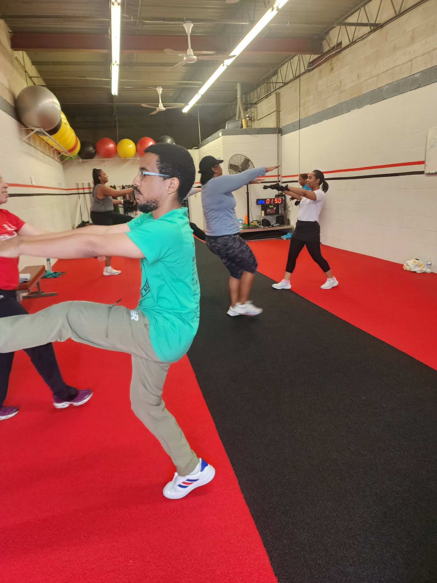 Person in teal shirt and beige pants stretches on red and black mat in gym, with others in background.