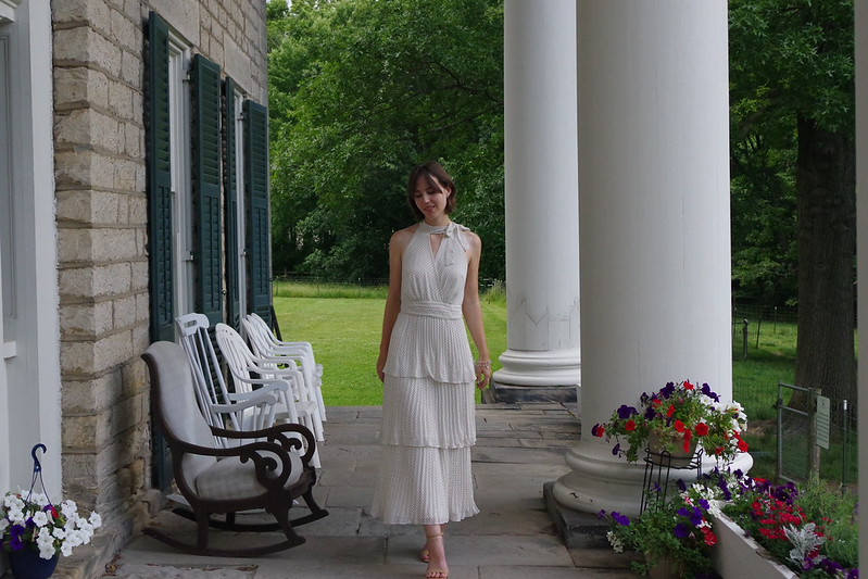 Woman in a white dress standing on a porch with columns, chairs, and flowers, greenery in the background.