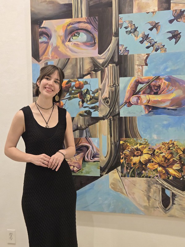 Young woman with short hair in a black dress smiling in front of colorful artwork with trees, birds, and flowers.