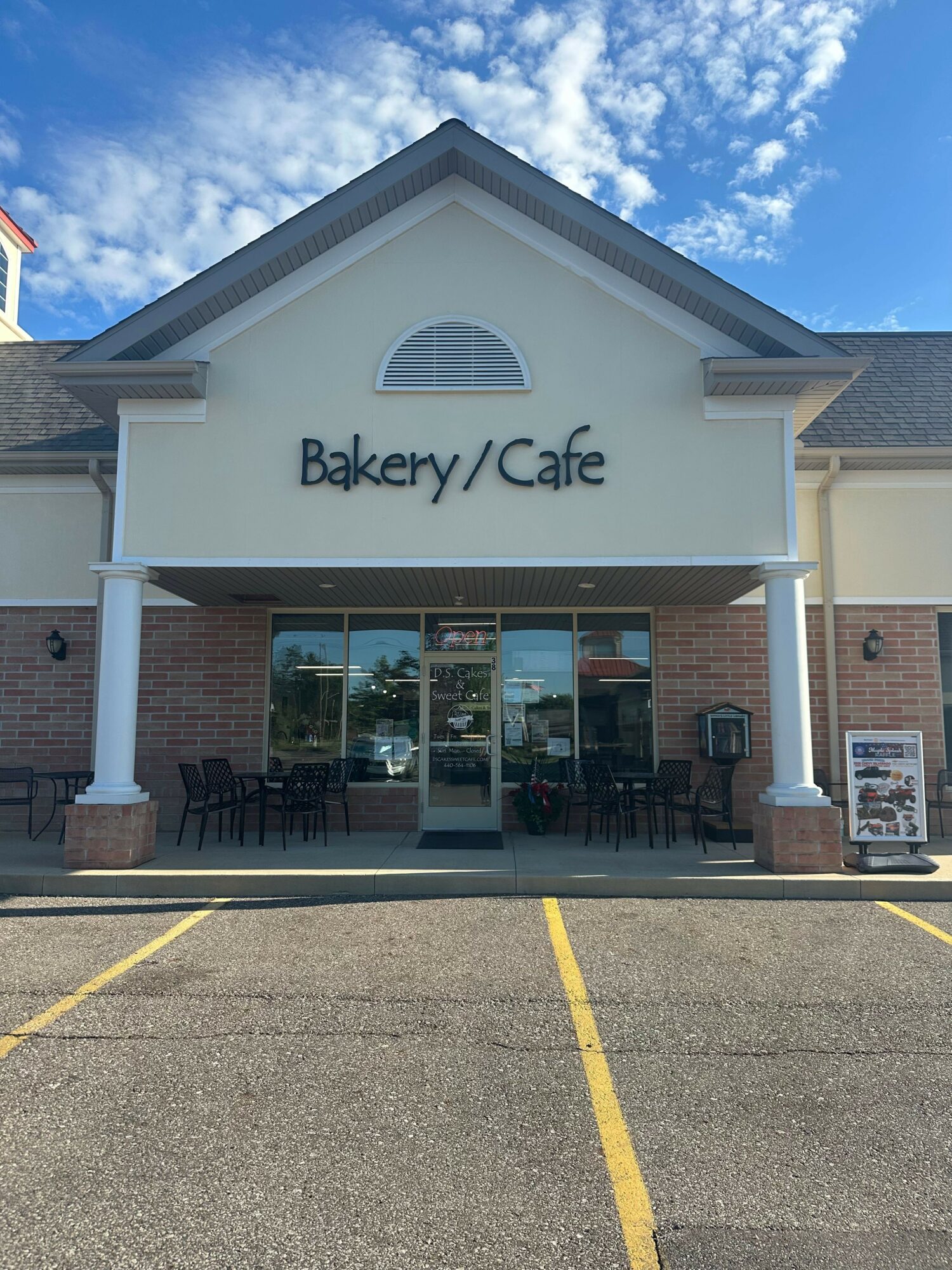 Bakery/Cafe storefront with large windows and a sign, brick exterior, and outdoor seating area, under a partly cloudy sky.