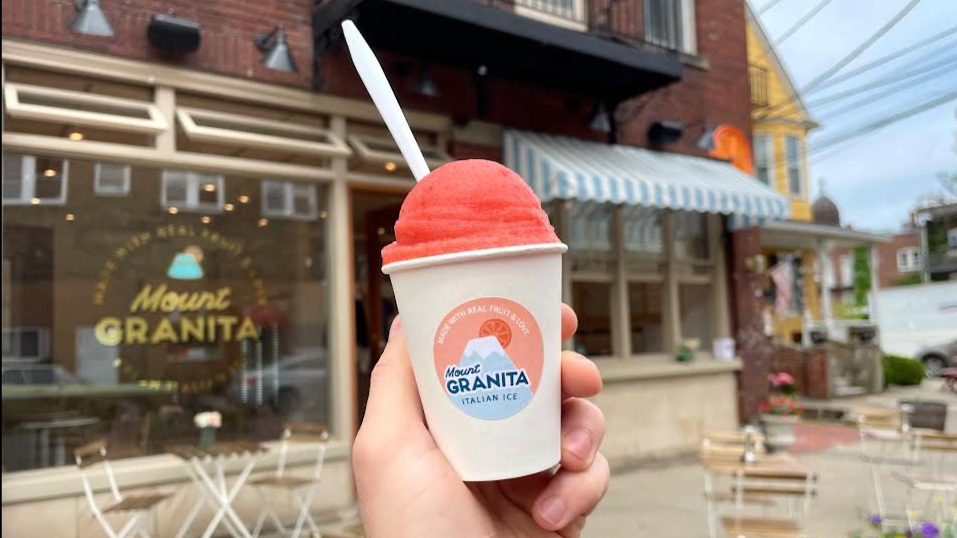 Hand holding a cup of pink ice cream with a straw in front of a building with a sign that reads 'Mount Granita'.