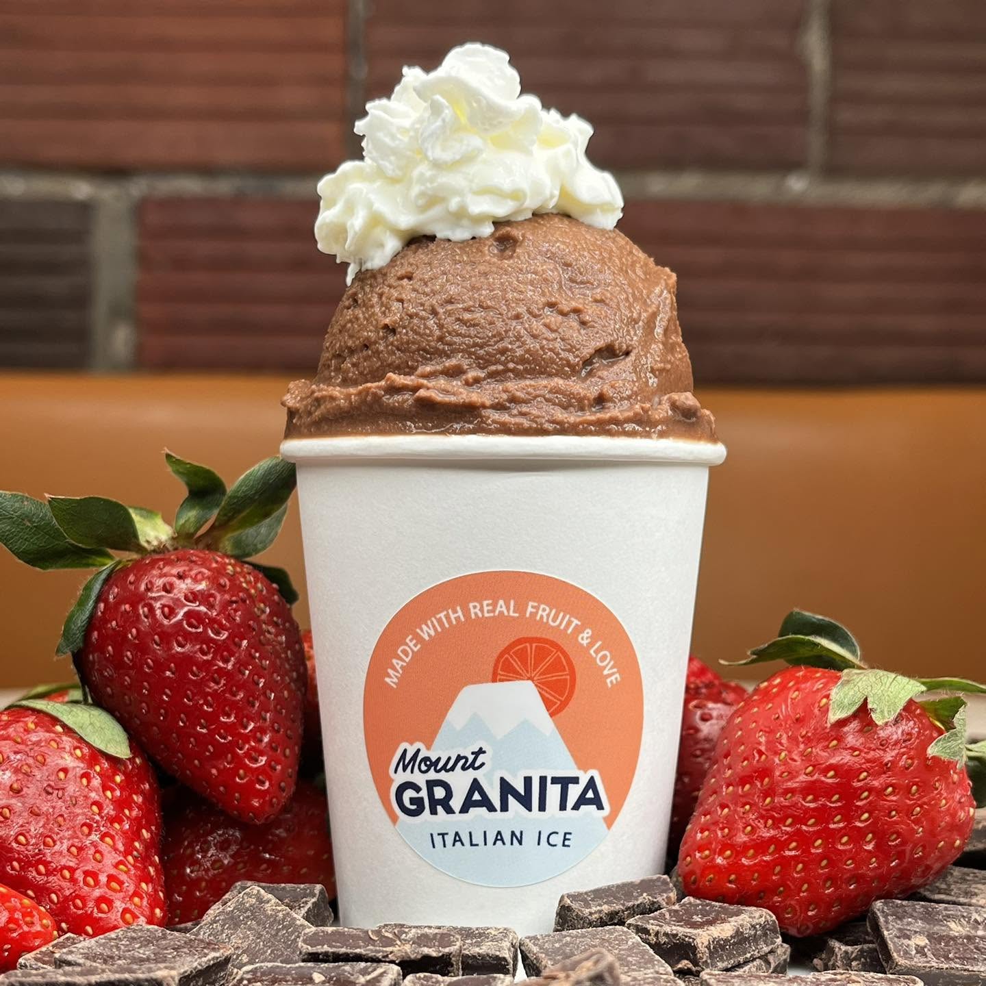 Ice cream in a cup with whipped cream, surrounded by strawberries and chocolate pieces, against a brick wall background.