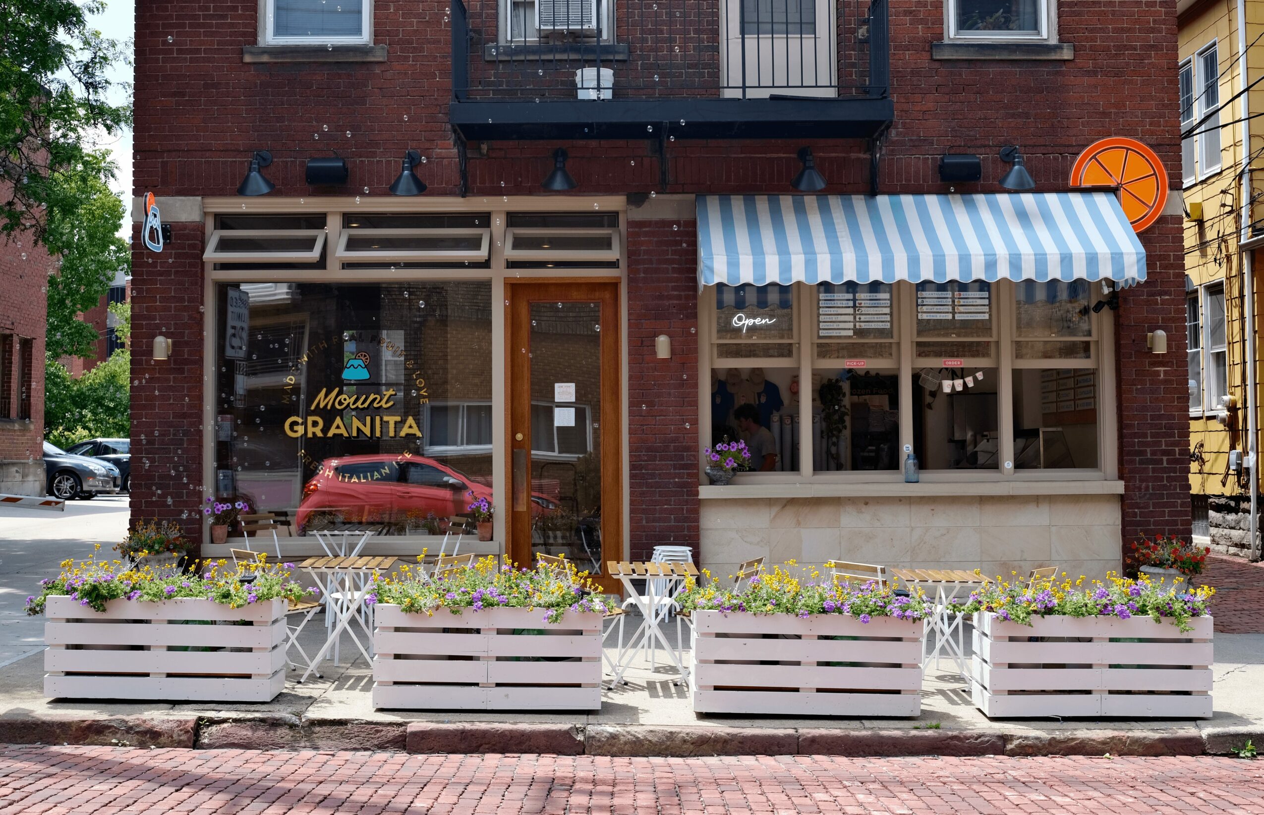 Storefront with large windows, striped awning, outdoor planters, and a red car reflected in the glass.