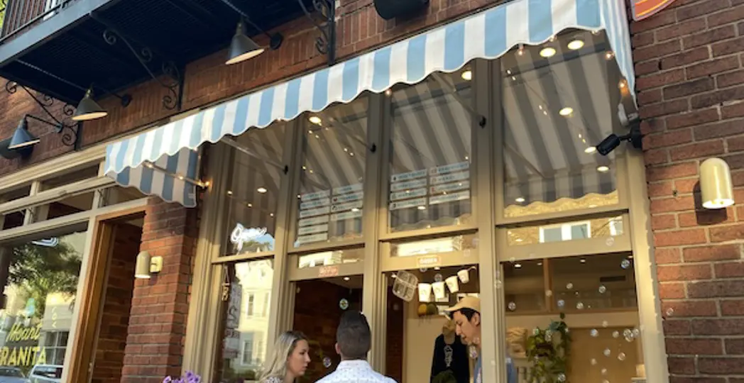People standing outside a storefront with a striped awning, brick building, and large glass windows.