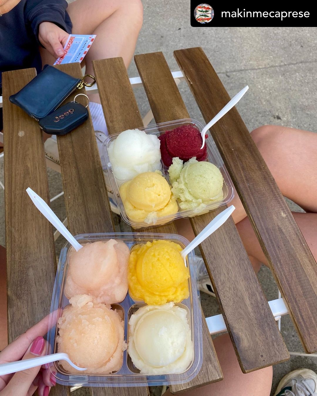 Two clear plastic containers with four scoops of ice cream on a wooden bench, with a person's hand holding a spoon and phone nearby.