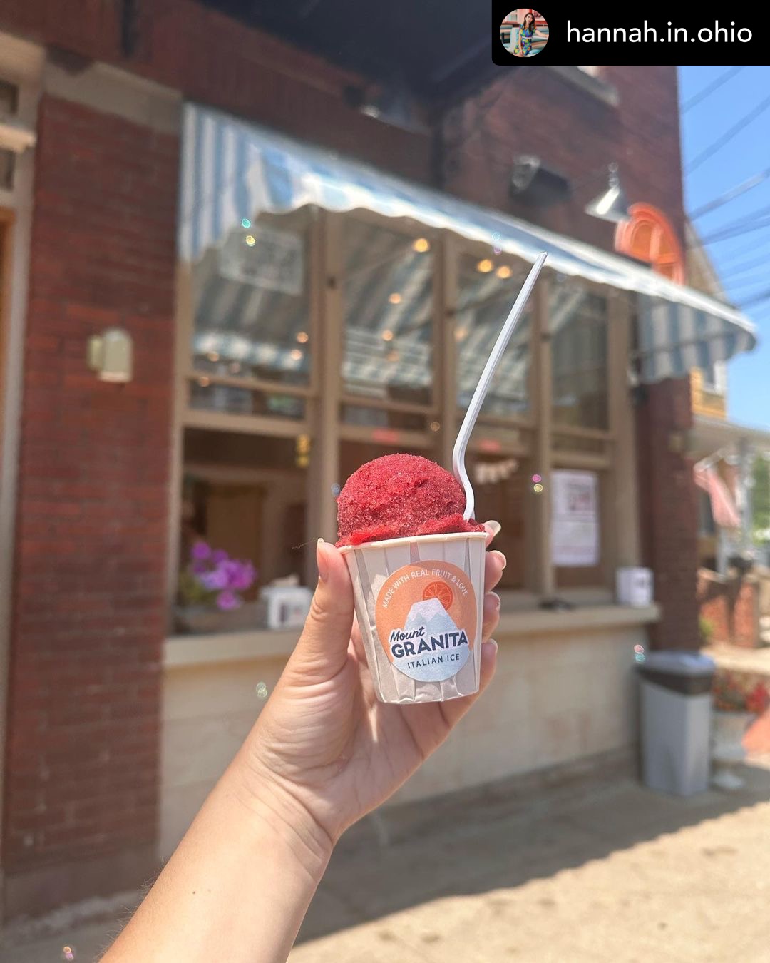 Hand holding a cup of red ice cream with a spoon outside a brick building with large windows and an awning.