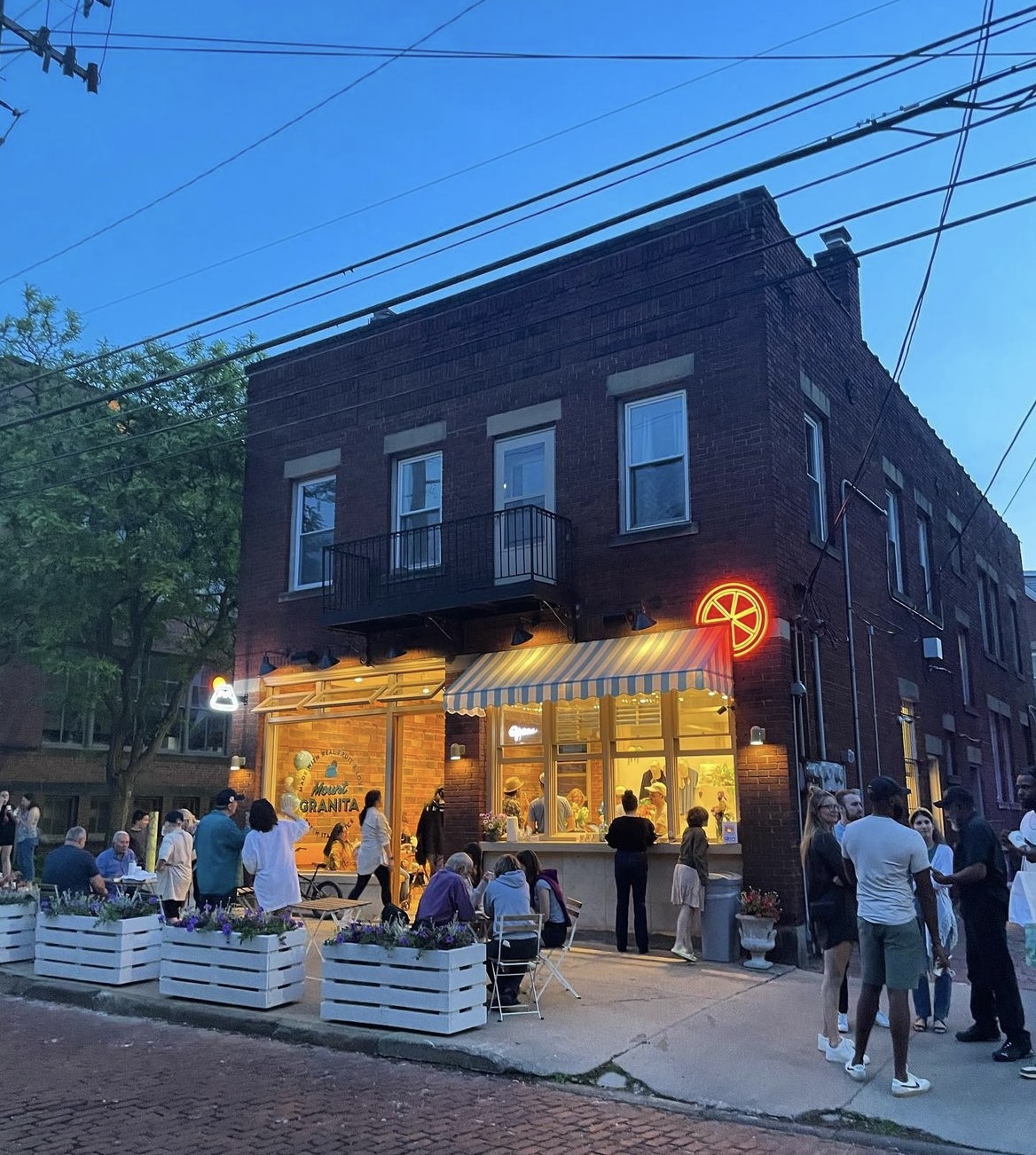 People gather outside a brick building with a yellow storefront and striped awning, illuminated by streetlights and a neon sign, during evening.