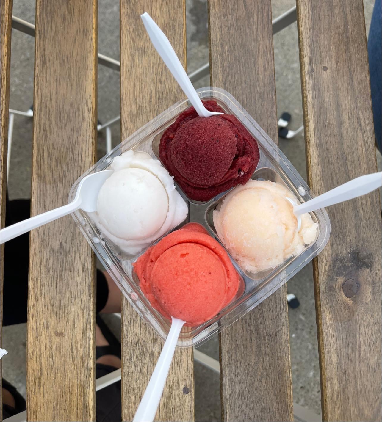 Four scoops of ice cream in a clear container on a wooden surface, with white plastic spoons, in red, white, peach, and pink colors.