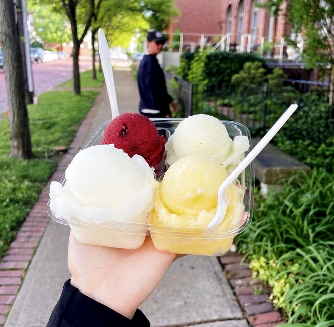 Hand holding a cup with three scoops of ice cream in a park, with a person walking in the background.
