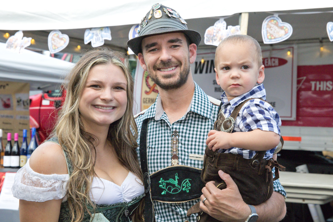 Family of three smiling at a fair, with a woman, man, and young boy, in front of a food stand.