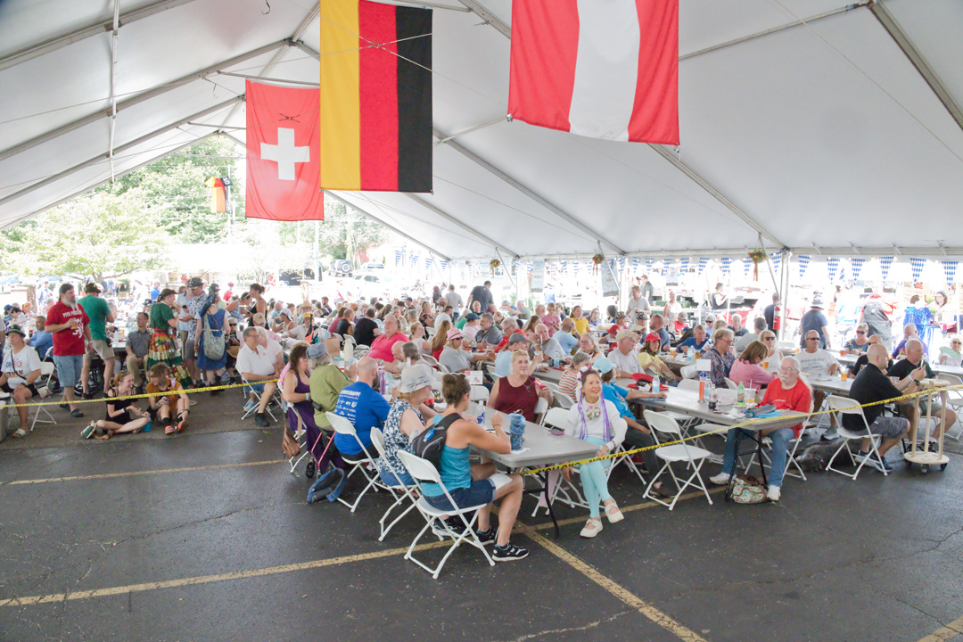 People seated at tables inside a large tent with flags hanging from the ceiling, some standing outside, bright daylight.