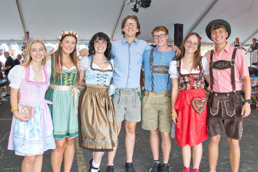 Group of seven young people smiling, standing together indoors, some wearing traditional Bavarian clothing, with a tent ceiling above.