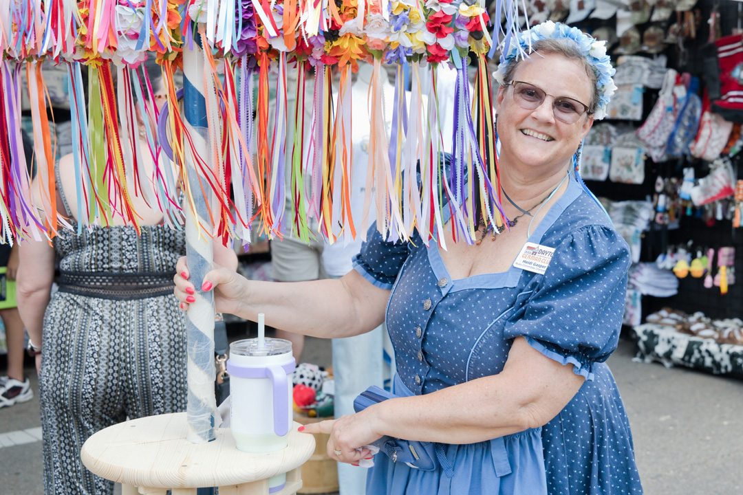 Smiling woman with glasses and blue dress at market stall with colorful ribbons, holding a bag, surrounded by other shoppers.
