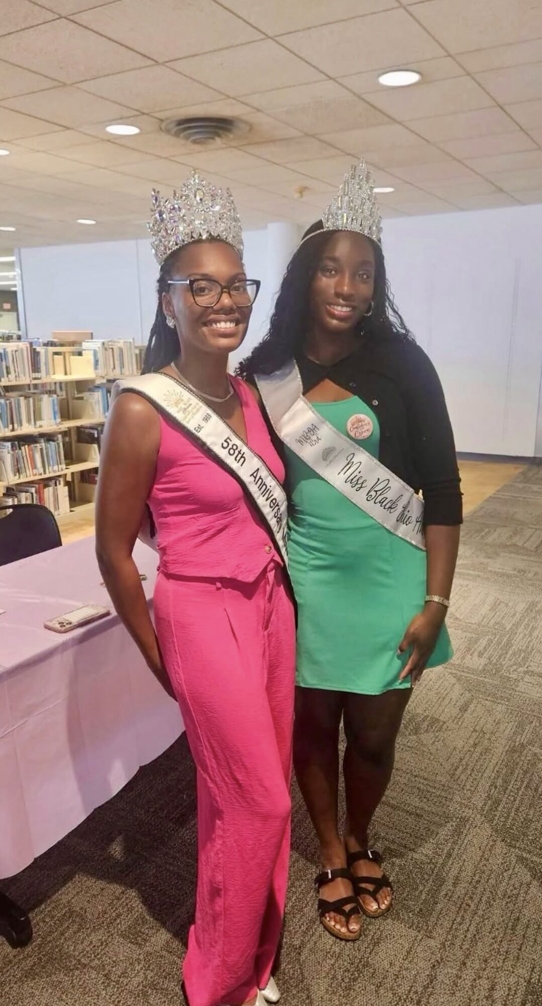 Two women wearing crowns and sashes, standing indoors with bookshelves in the background.