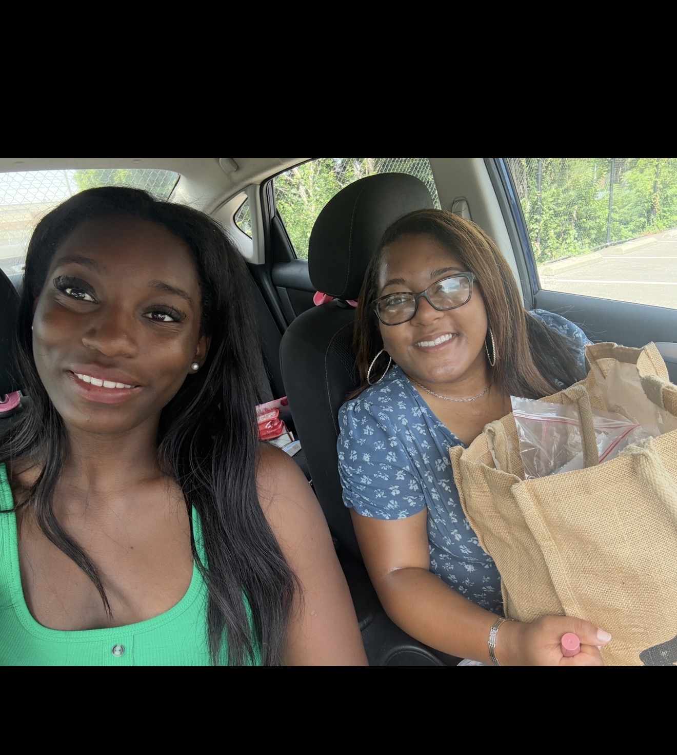 Two women sitting in a car, smiling, with a paper bag on the passenger side. Outside greenery visible through windows.