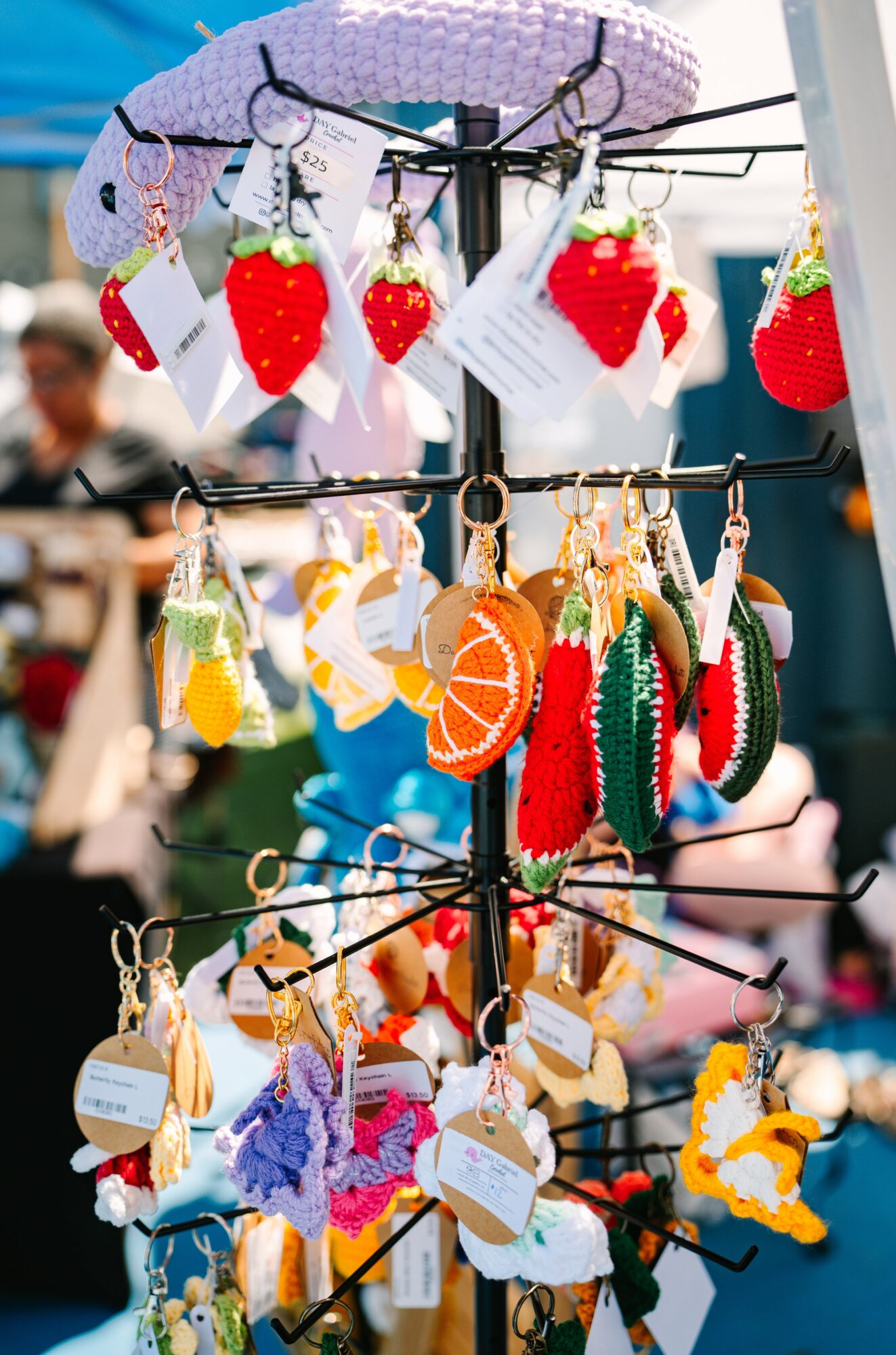 Display of colorful handmade strawberry and fruit-shaped ornaments hanging on a stand at an outdoor market.