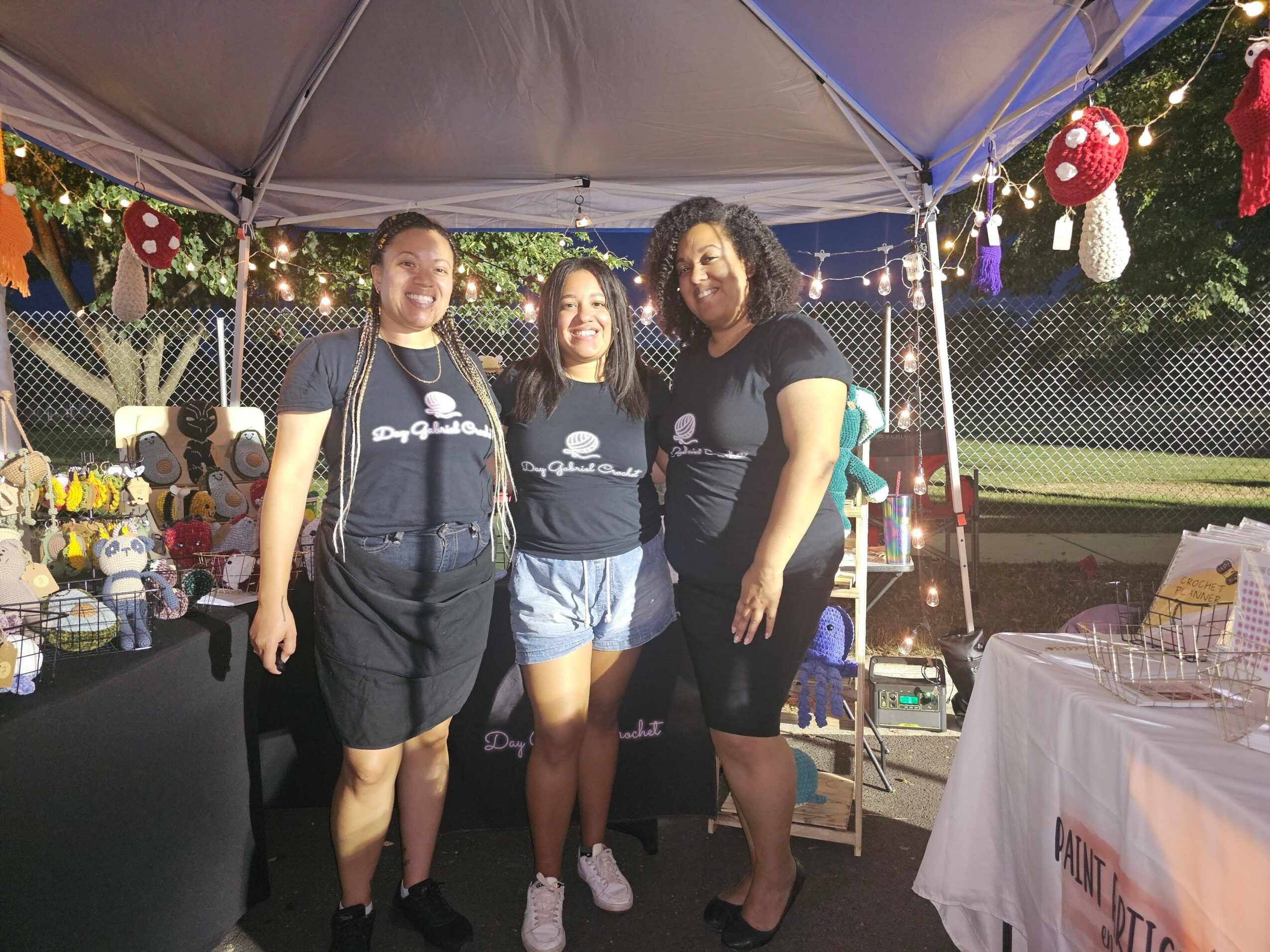 Three women standing under a canopy at an outdoor market or fair, with various items on tables behind them.