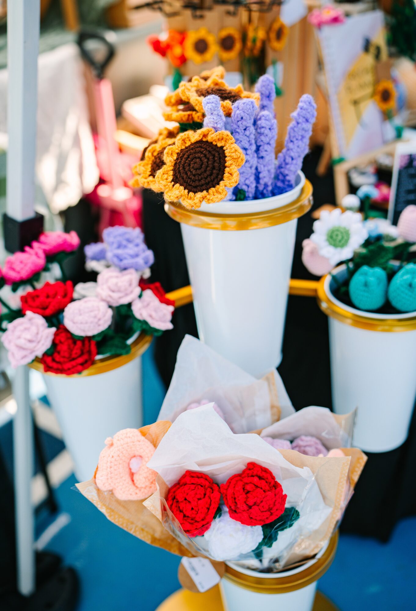 Colorful handmade crochet flowers in white vases at an outdoor market stall.