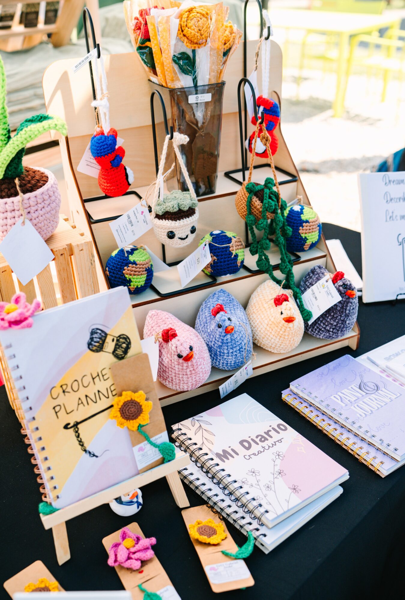 Display of handmade crochet items, notebooks, and decorative flowers on a table outdoors, with a yellow railing in the background.