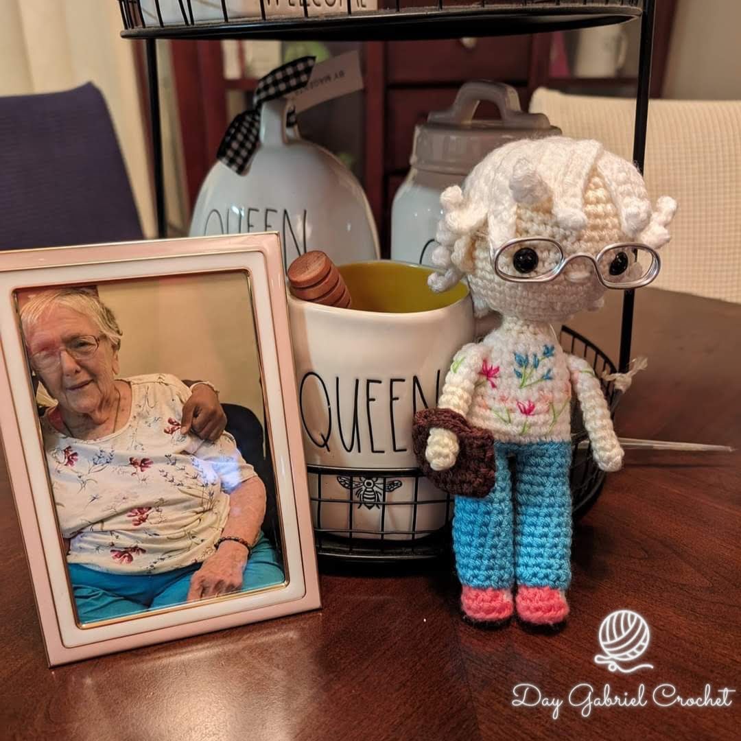 Photograph of an elderly woman in a floral top, smiling, with a knitted doll wearing glasses and colorful clothes nearby, on a table.