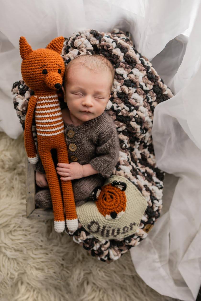Baby sleeping on a blanket with a knitted fox toy, surrounded by soft fabrics and a plush rug.