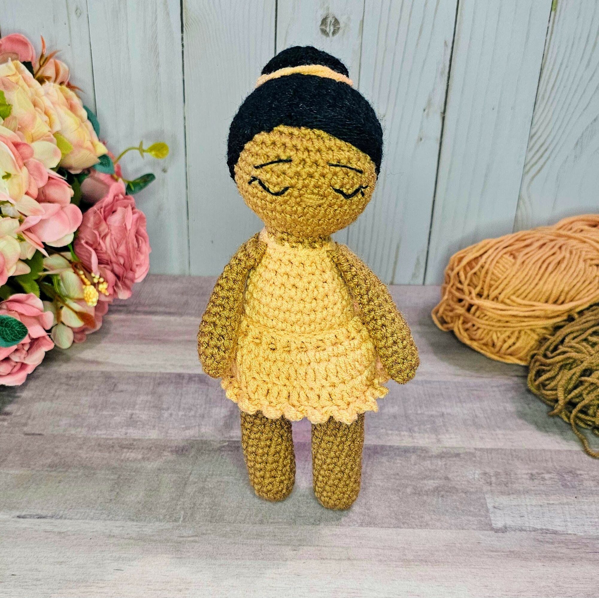 Handmade crocheted doll with black hair, yellow dress, and closed eyes, standing on a wooden surface with flowers and yarn nearby.