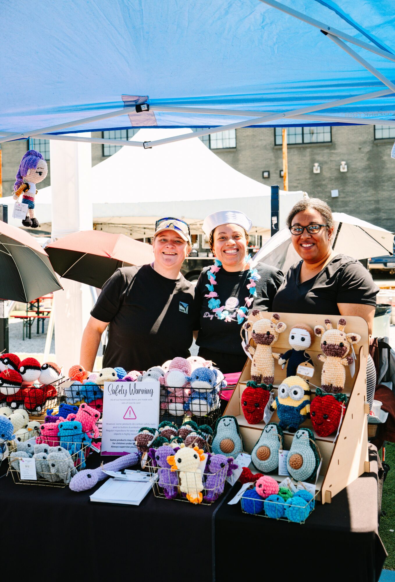 Three women smiling behind a table with colorful handmade plush toys at an outdoor market stall.