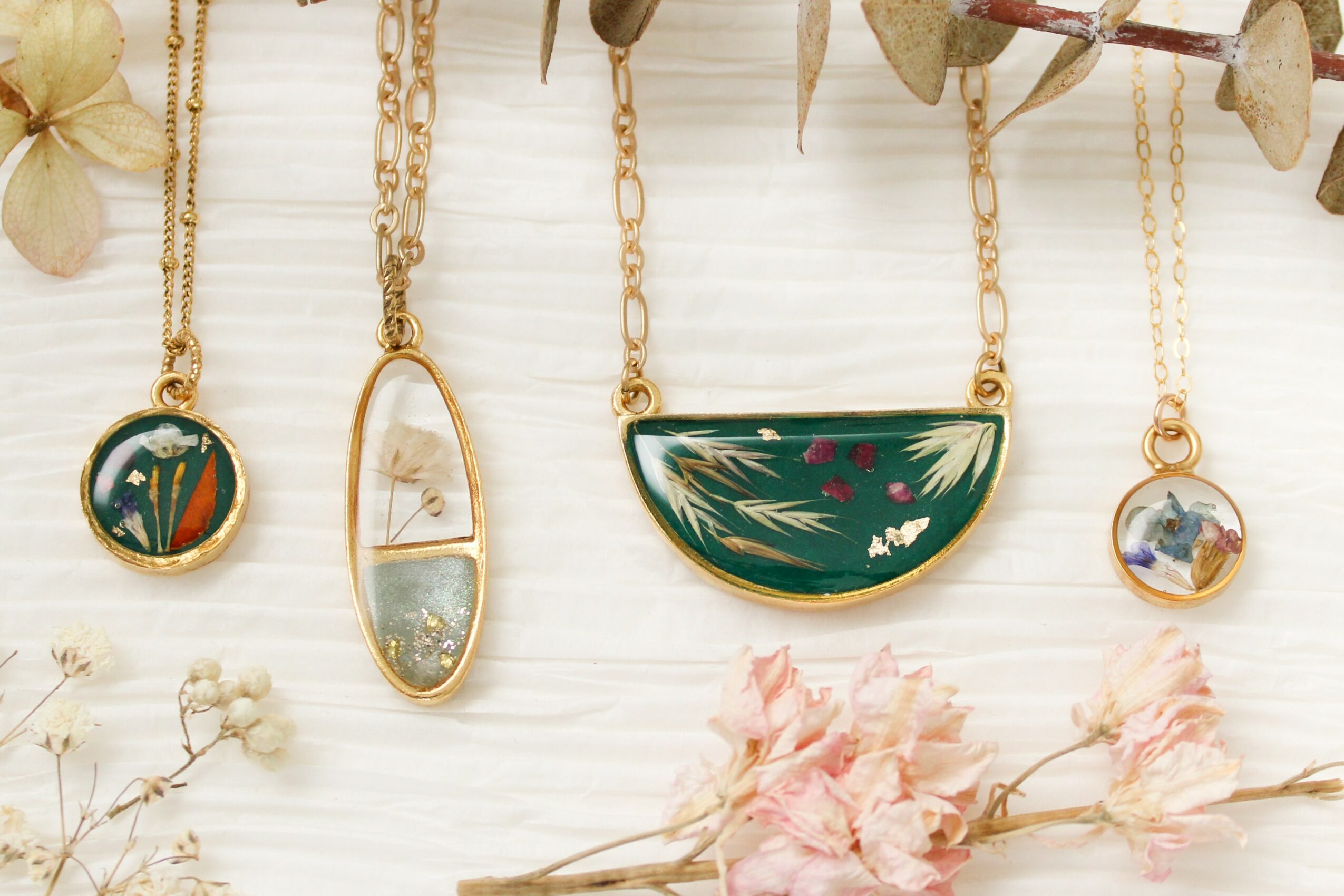 Four necklaces with colorful pendants hanging against a white background, decorated with dried flowers and leaves.