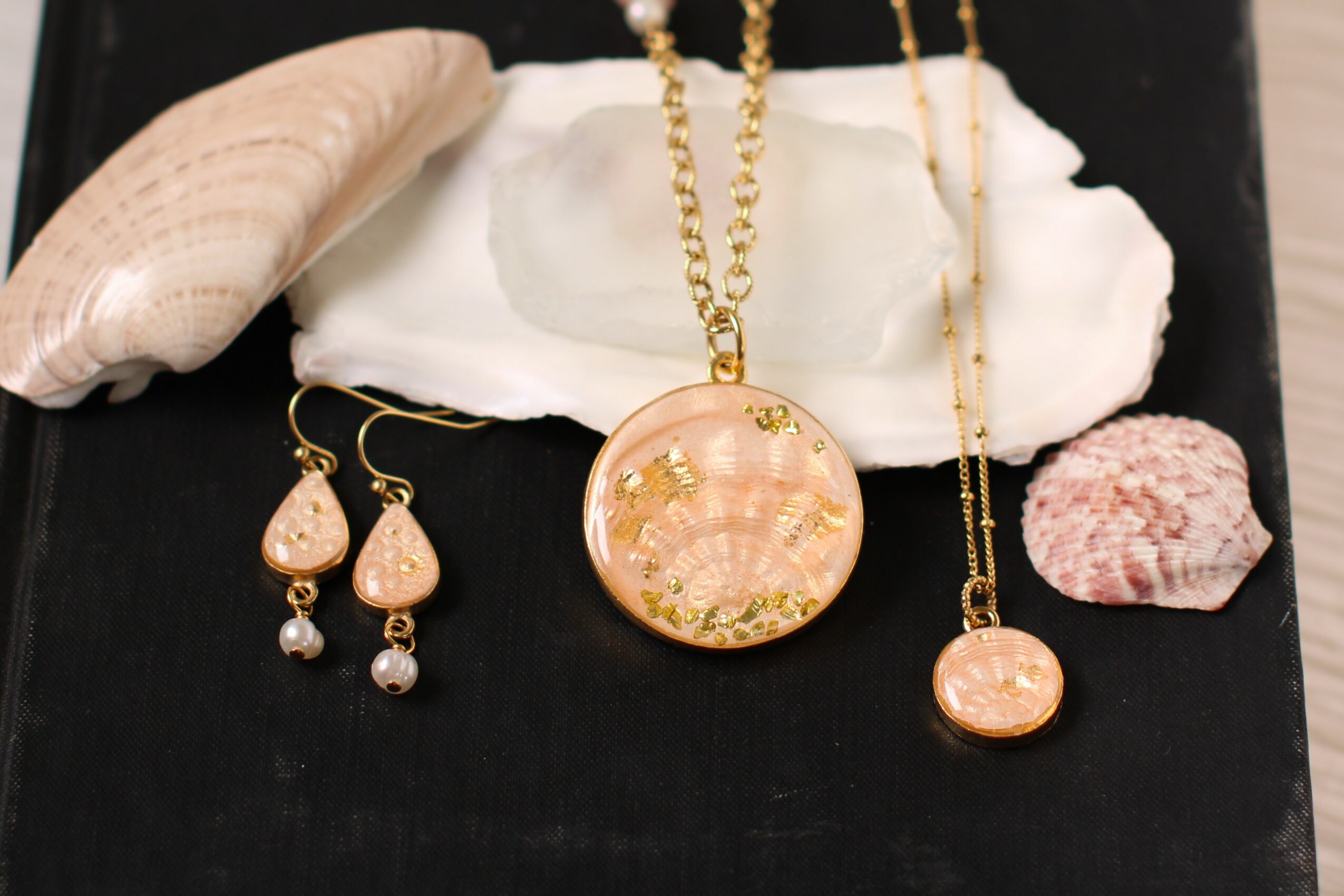 Jewelry including a gold necklace with a round pendant, matching earrings, and a smaller pendant, displayed with seashells and coral on black background.