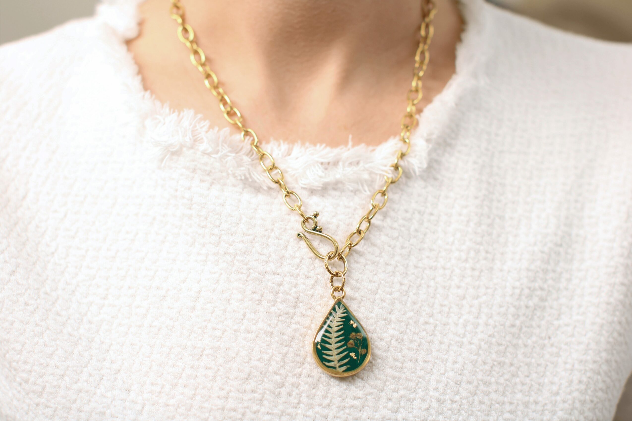 Person wearing a gold chain necklace with a green teardrop pendant, dressed in a white textured top.