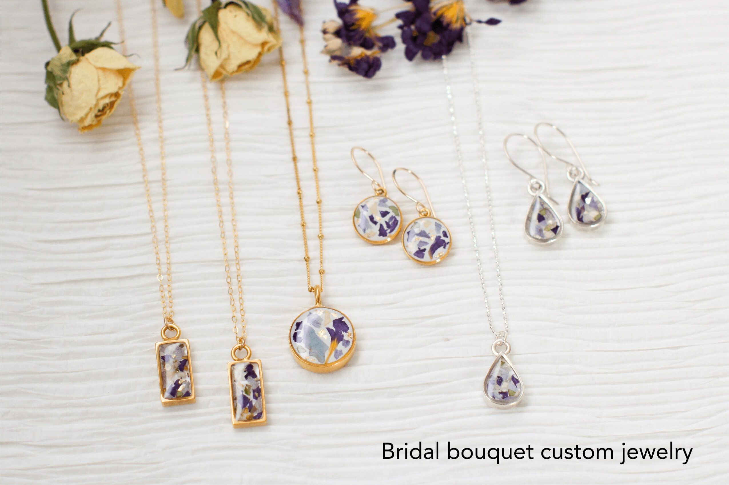 Two necklaces and two pairs of earrings with purple and silver designs on a white surface, with dried flowers nearby.