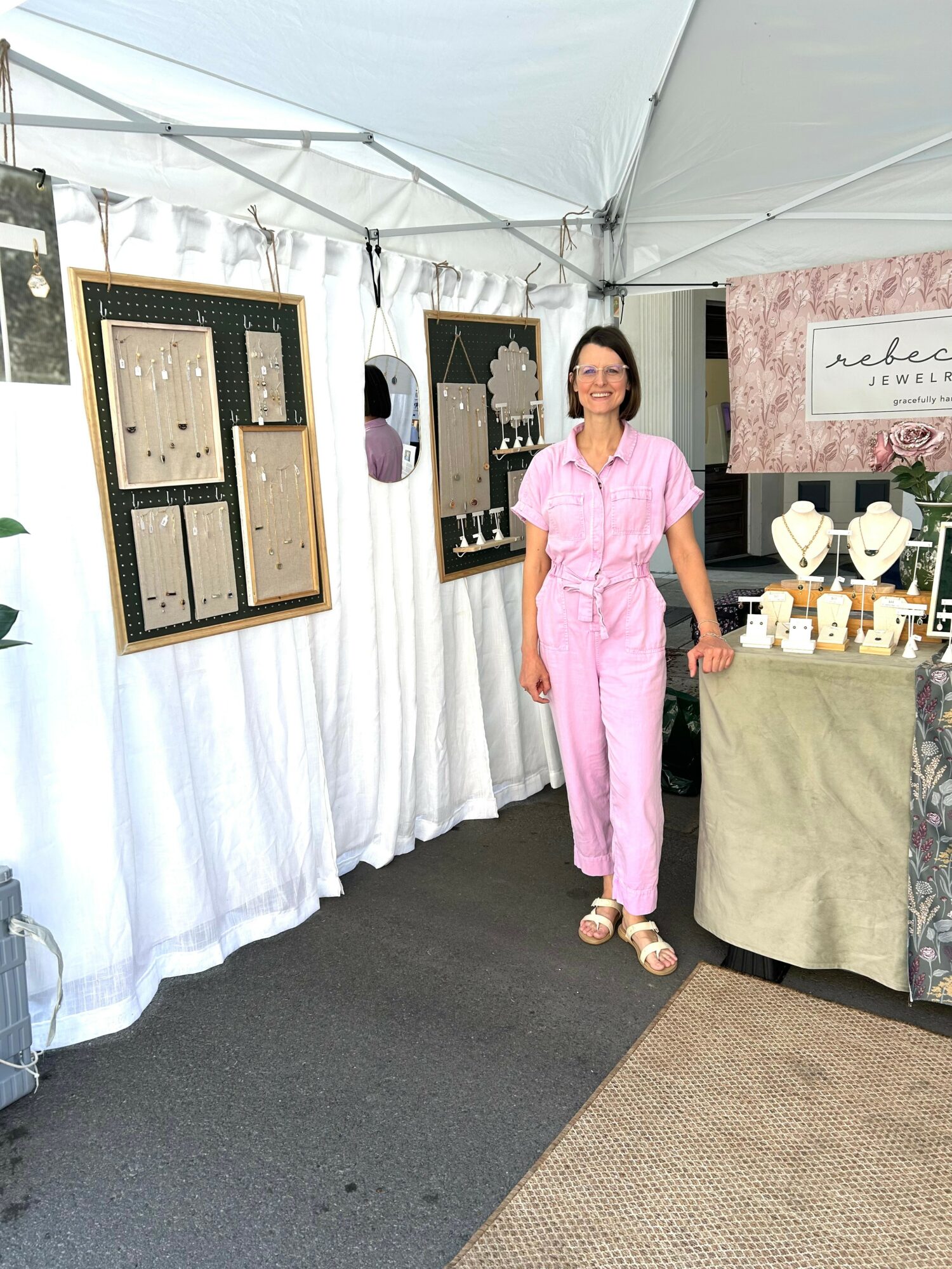 Woman in pink jumpsuit standing inside a booth with jewelry displays and artwork, under a white canopy tent.
