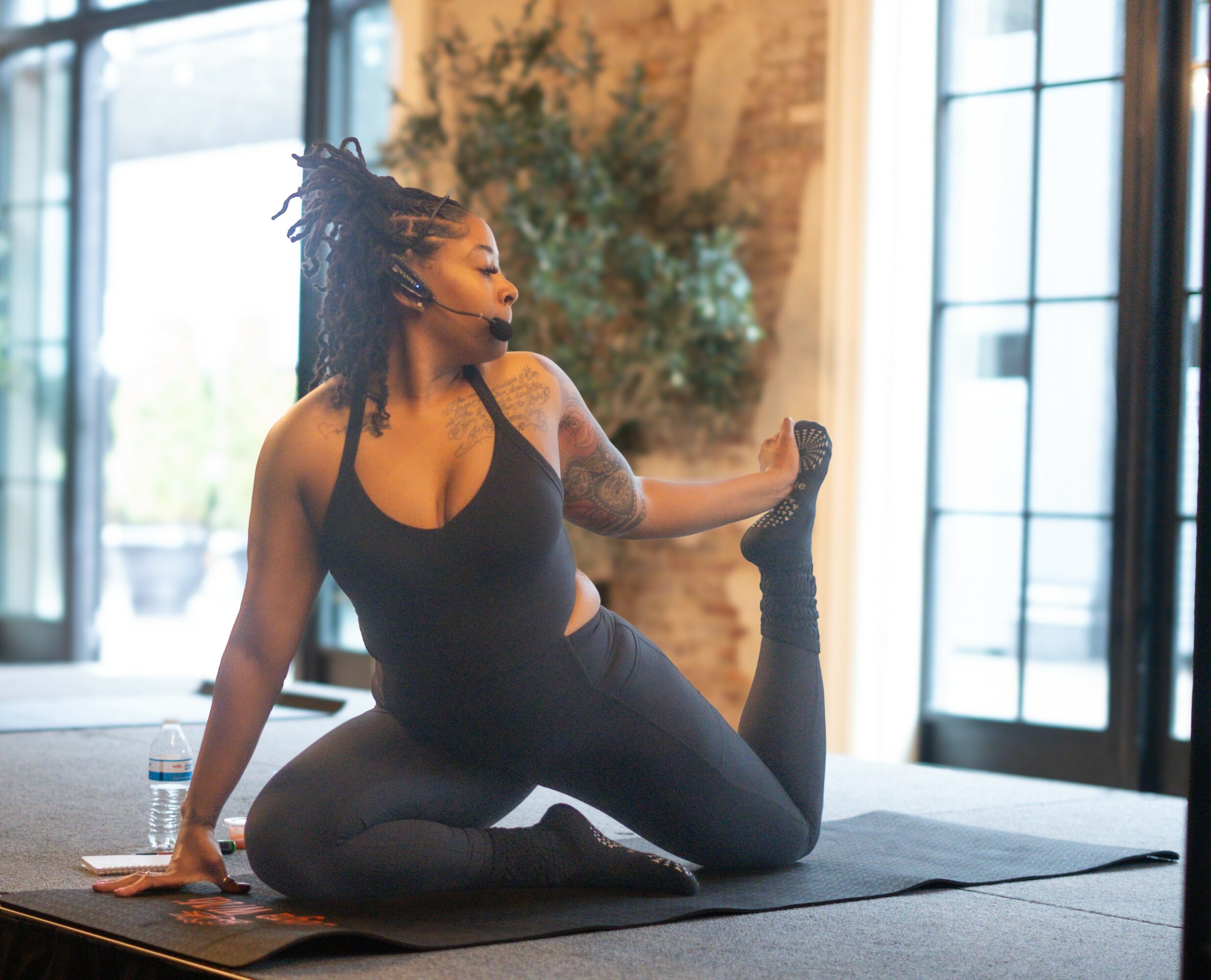Woman practicing yoga indoors on a mat, stretching her leg behind her, with large windows and plants in the background.