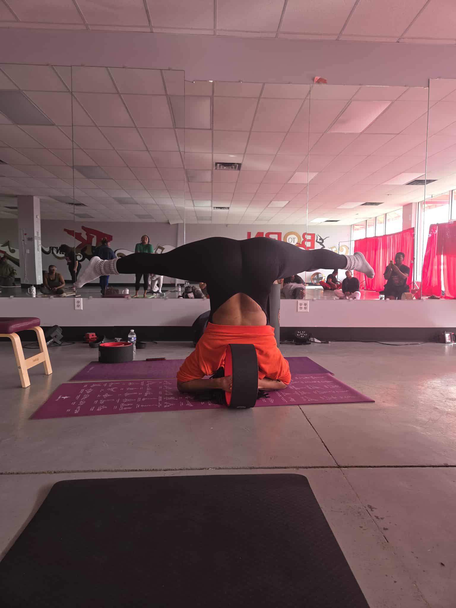 Person performing a headstand on a yoga mat in a fitness studio with mirrors and red curtains.