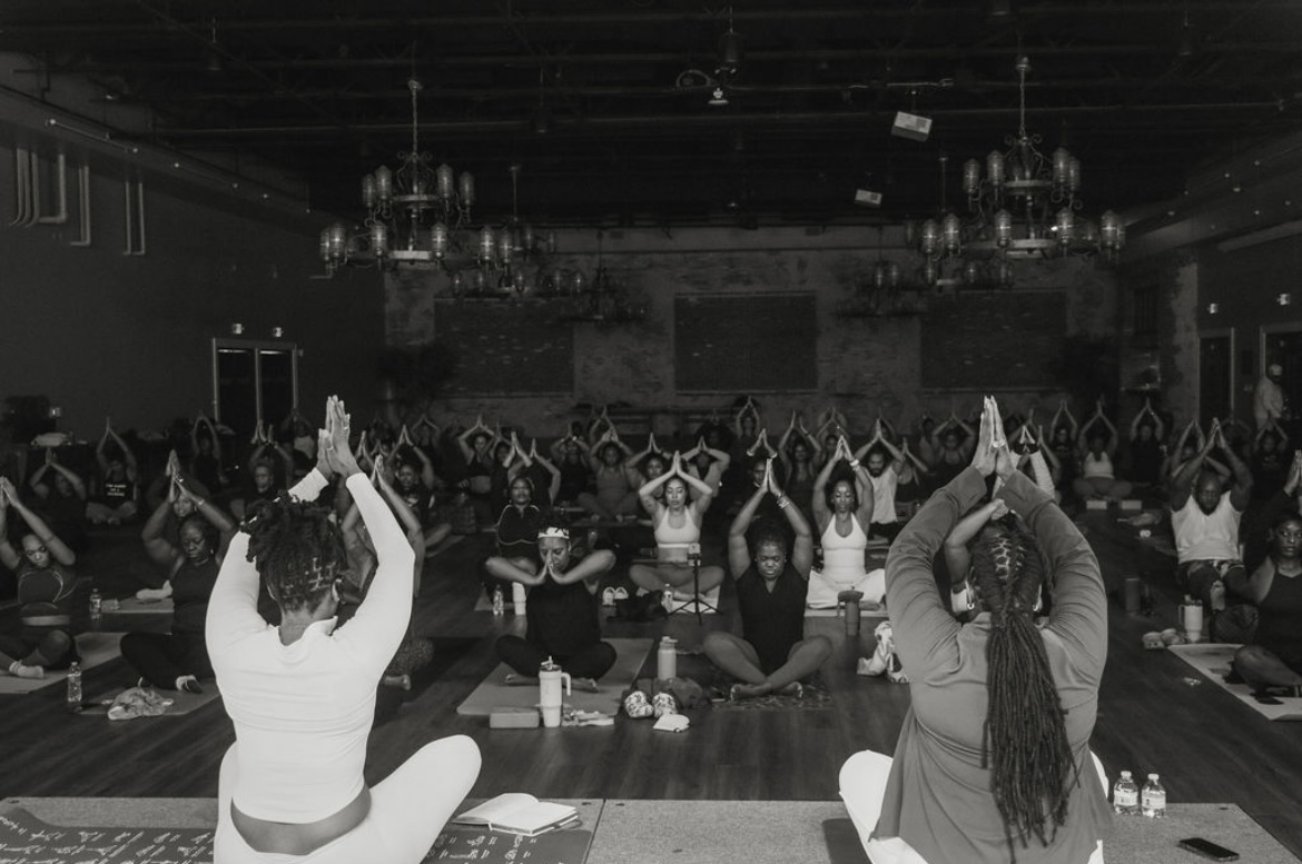People participating in a group yoga or meditation session in a large indoor hall, sitting cross-legged with hands raised above heads.