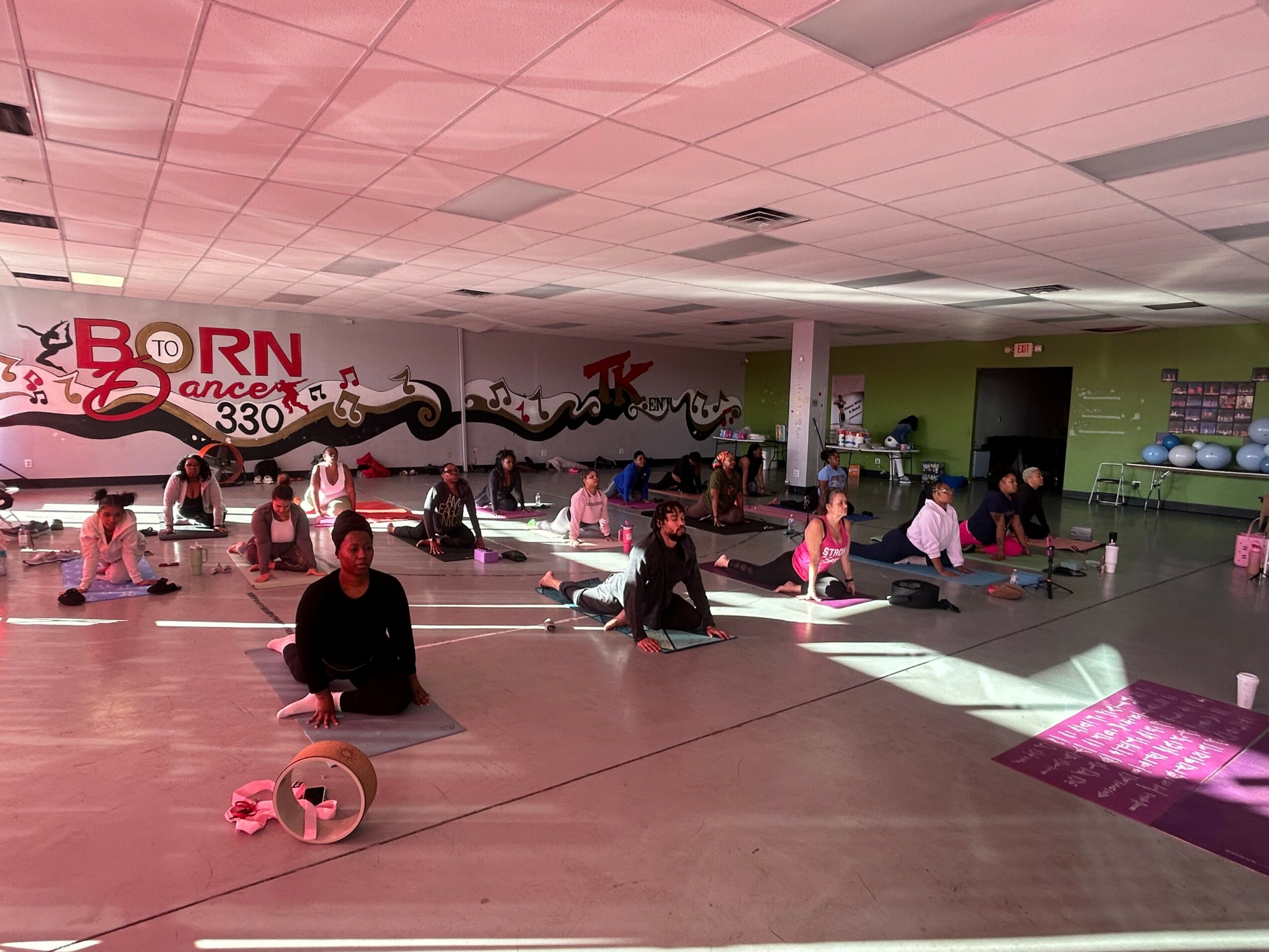 People practicing yoga in a fitness studio with pink and purple mats, weights, and a wall mural, sunlight streaming in.