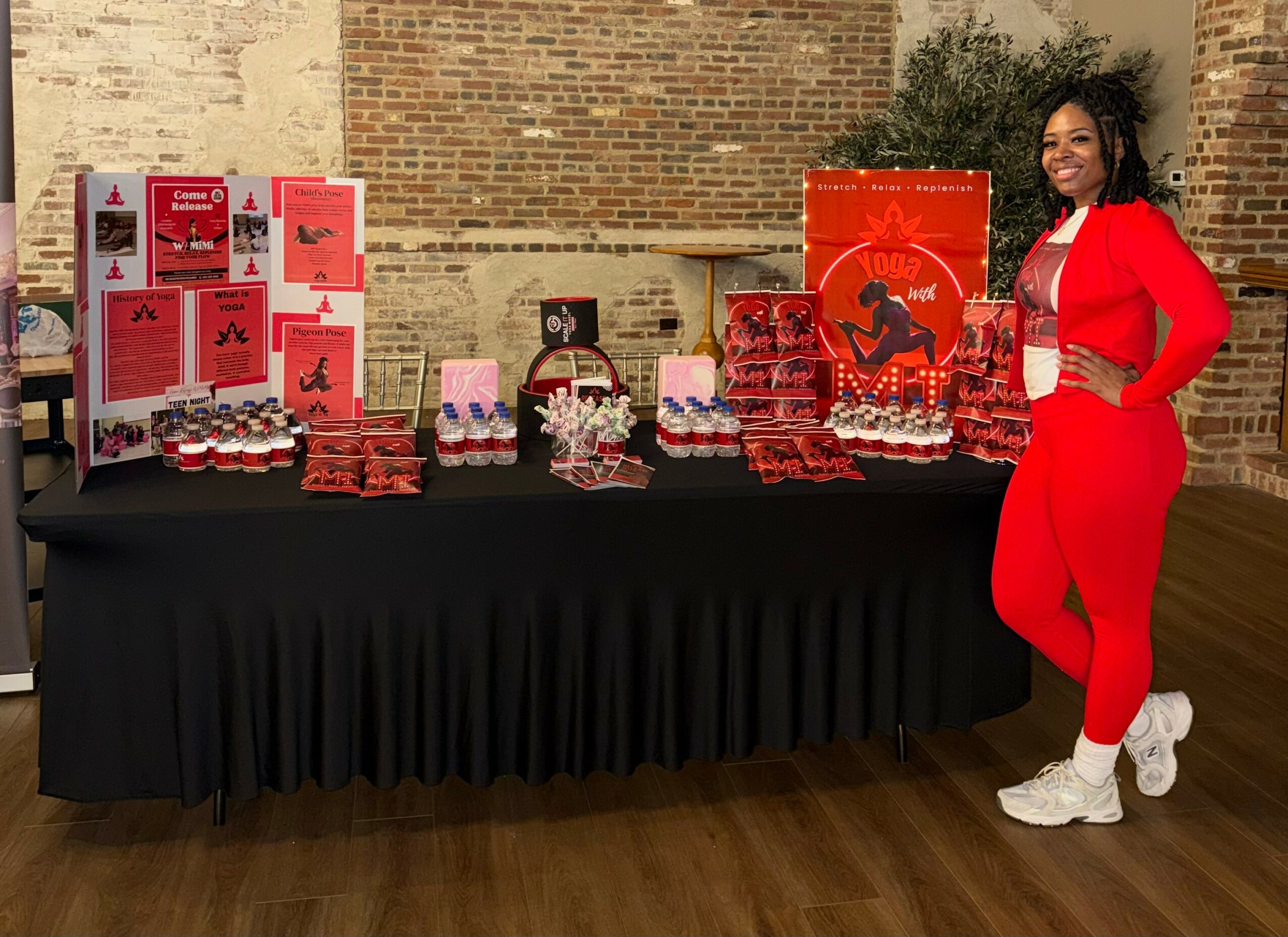 Woman in red outfit standing next to a table with red-themed items and decorations, brick wall background.